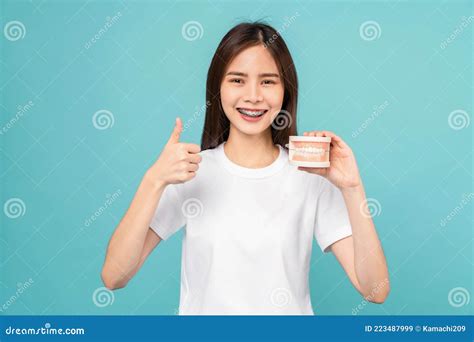 Woman Wearing Braces Showing Thumbs Up Or Like And Holding Tooth Model On Blue Background Stock