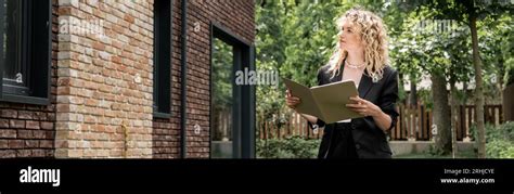 Attractive Blonde Real Estate Agent With Folder Looking At Building On Urban Street Banner