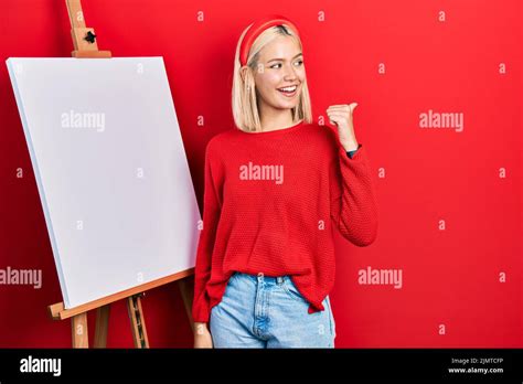 Beautiful Blonde Woman Standing By Painter Easel Stand Pointing Thumb Up To The Side Smiling