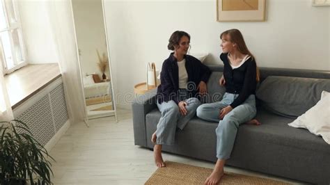 Two Girlfriends Or Sisters Talking And Laughing At Home On The Couch