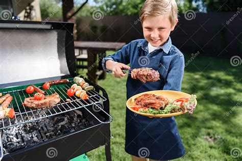 Kid Boy In Apron Preparing Tasty Stakes On Barbecue Grill Outdoors