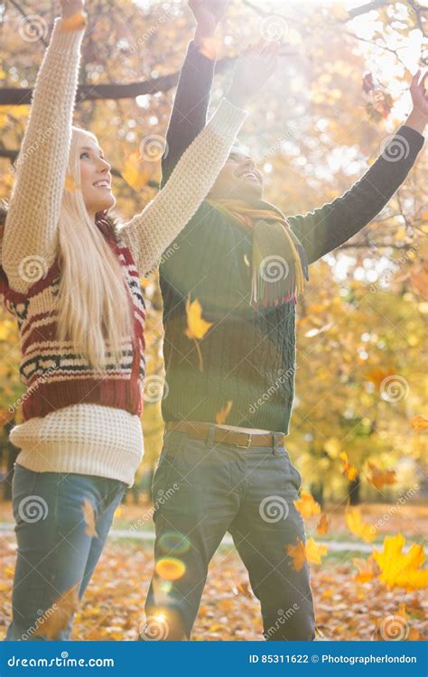 Happy Young Couple With Arms Raised Enjoying Falling Autumn Leaves In