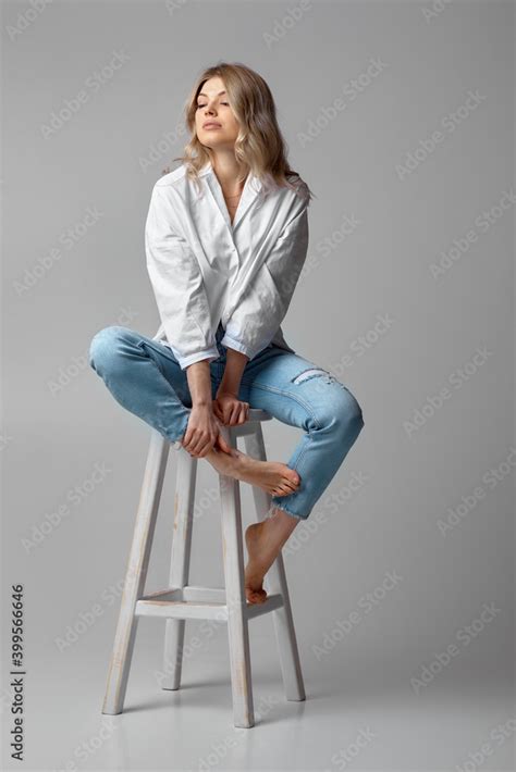 Beautiful Blonde Woman With Curly Hair In Jeans And A White Shirt Sitting On Chair On Studio