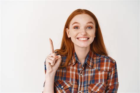 쾌활한 빨간 머리 여성이 손가락을 가리키고 하얀 치아로 웃고 광고를 보여주고 흰 벽 위에 서 있다 프리미엄 사진