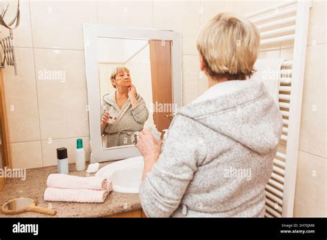 Senior Woman In Bathrobe Touching Her Soft Face Skin Looking In Mirror