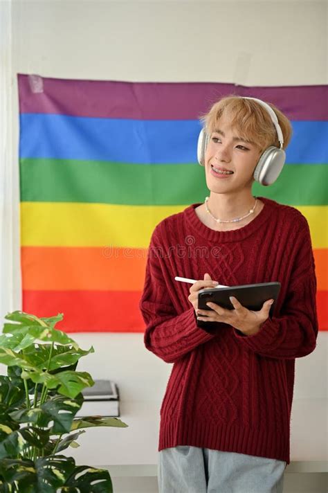 A Happy Asian Gay Man Is Listening To Music While Standing In His Room With An Lgbt Flag Behind