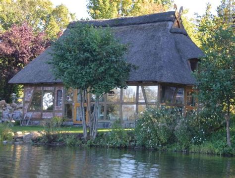 A House With A Thatched Roof Sitting On The Side Of A Lake Next To Trees