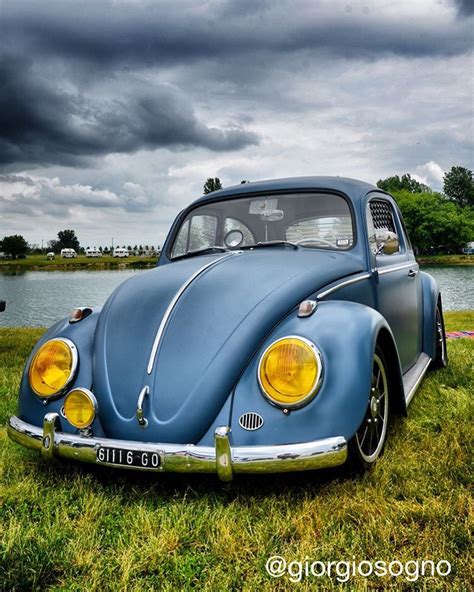 an old blue vw bug is parked in the grass near some water and clouds