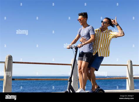 Happy Biracial Gay Male Couple Having Fun Riding Scooter Together On Promenade By Sea Copy