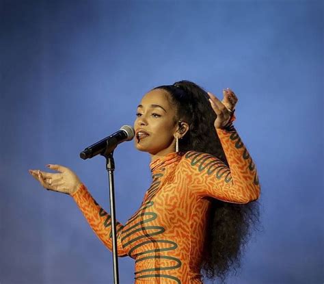 A Woman In An Orange And Green Bodysuit Holding A Microphone Up To Her