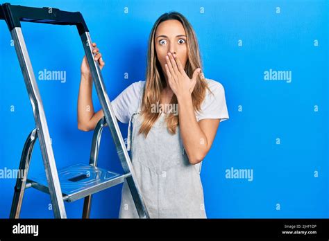 Beautiful Hispanic Woman Standing By Ladder Covering Mouth With Hand