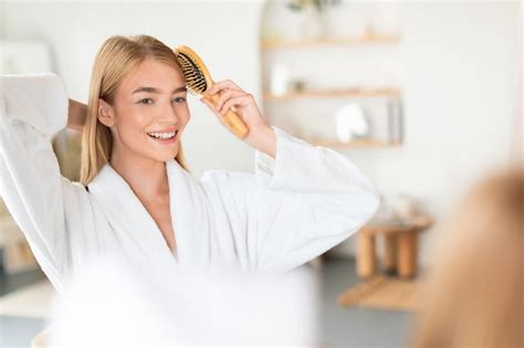 Premium Photo Attractive Blonde Woman Combing Hair With Wooden Brush In Bathroom