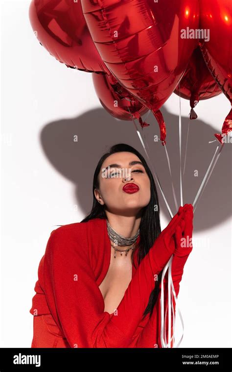 Stylish Brunette Woman In Red Gloves Holding Heart Shaped Balloons And Blowing Air Kiss On White