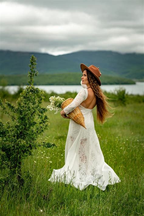 A Middle Aged Woman In A White Dress And Brown Hat Stands With Her Back