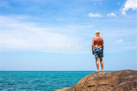 Mature Blond Man With Bare Torso Stands On A Rock On The Seashore And Looks Into The Distance