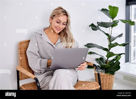 Excited Caucasian Blonde Woman In Glasses Sitting On Chair Using