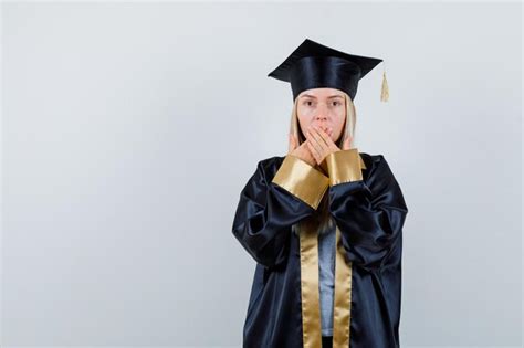 Fille blonde en robe de graduation et casquette couvrant la bouche avec les mains et à la