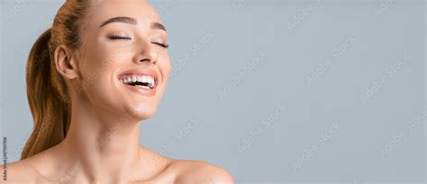 Woman With Long Blonde Hair Pulled Back In A Ponytail Smiling With Her Eyes Closed Against A