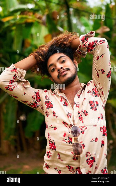 Confident Gay Man Of Color Poses With Flair In Floral Shirt Amidst Plants Hands Rest On Head