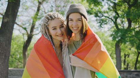 Happy Lesbian Couple Hugging And Holding Rainbow Flag In Park And Looking At The Camera At