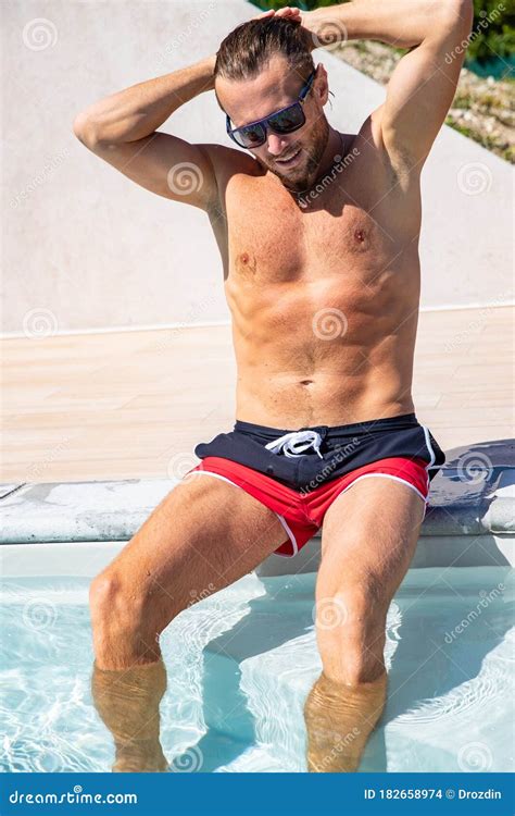 The Handsome Brutal Man In Sunglasses With A Long Hair And Naked Torso Sits Near The Pool