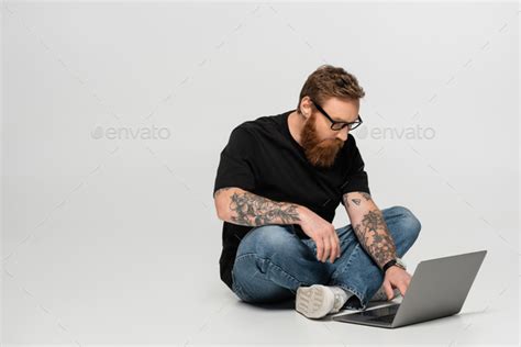 Concentrated Bearded Man In Eyeglasses Using Laptop While Sitting With