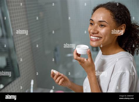 Smiling African Woman Applying Moisturizing Cream Standing In Home