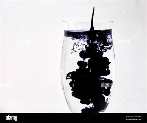 Blue And Black Ink Dropping Into A Wine Glass Of Water On A White