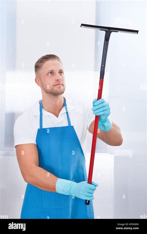 Male Janitor Using A Squeegee To Clean A Window In An Office Wearing An