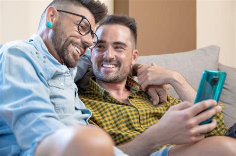 Alegre pareja gay sentada en el sofá abrazándose y mirando el teléfono móvil viendo una película