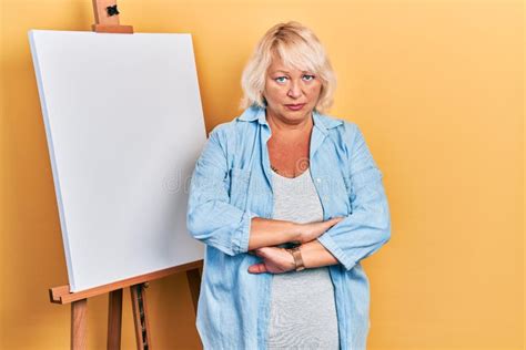 Middle Age Blonde Woman Standing By Painter Easel Stand Skeptic And