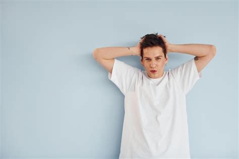 Joven gay está parado en el estudio y posando para una cámara con una camisa blanca casual