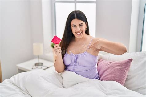 Jovem Morena Vestindo Lingerie E Segurando Preservativo Na Cama Apontando O Dedo Para Um