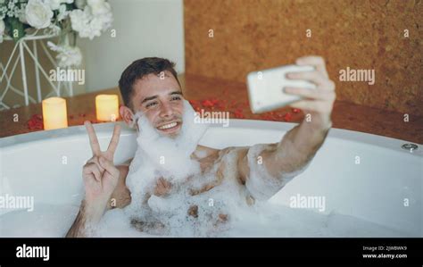 Handsome Cheerful Guy Is Taking Selfie In Bathtub With Soap Foam On His Beard Using Smart Phone