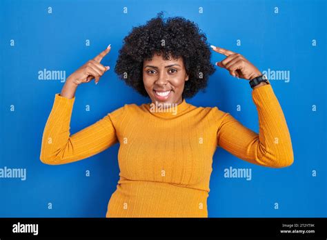 Black Woman With Curly Hair Standing Over Blue Background Smiling Pointing To Head With Both