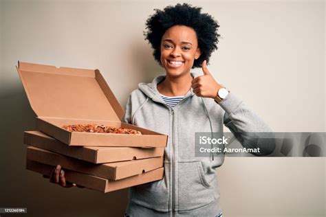 젊은 아프리카계 미국인 Afro 여자 곱슬 머리 들고 배달 상자 와 이탈리아 피자 행복 와 큰 미소 하 고 확인 기호 엄지