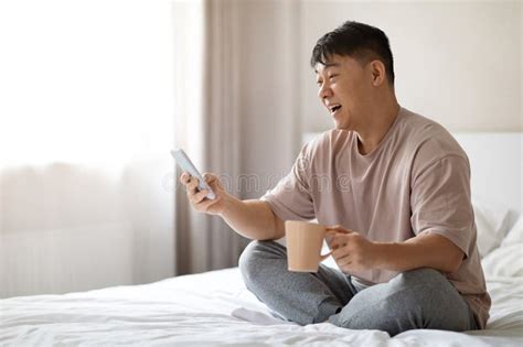 Positive Korean Man Using Smartphone Sitting On Bed At Home Stock