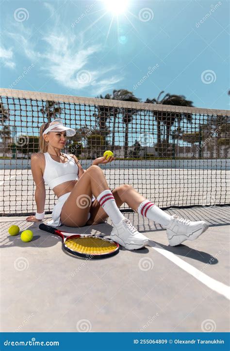 Beautiful Girl Sitting With The Ball On The Tennis Court In A Tennis Outfit In A Short Skirt