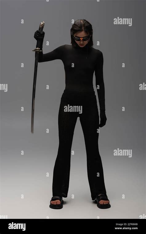 Full Length Of Brunette Asian Woman In Sunglasses And Black Outfit Holding Katana On Grey Stock