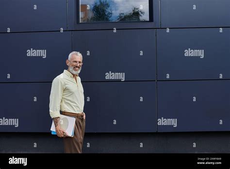 A Mature Gay Man With A Beard Smiles As He Stands Outdoors Near A Modern Building Holding Books