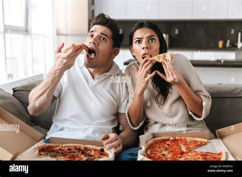 Cheerful young couple sitting on a couch at home, eating pizza ...