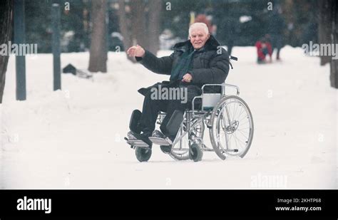 An Old Man Veteran Sitting In A Wheelchair And Waving With His Hand To