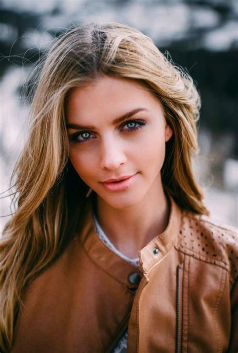 A Woman With Long Blonde Hair And Blue Eyes Wearing A Brown Leather Jacket Looking At The Camera