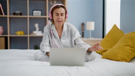 Middle Age Hispanic Woman Listening To Music Sitting On Bed At Bedroom