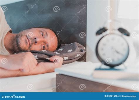 Sleepless Man Lying In Bed And Looking At Alarm Clock On Bedside