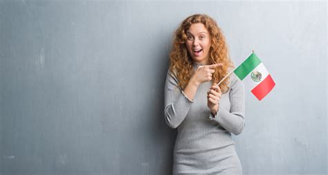 Young Redhead Woman Over Grey Grunge Wall Holding Flag Of Mexico Very