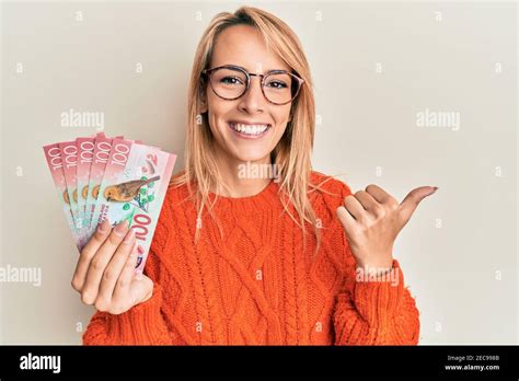 Beautiful Blonde Woman Holding 100 New Zealand Dollars Banknote Pointing Thumb Up To The Side