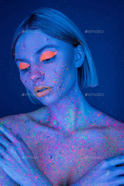 Nude Woman In Bright Neon Makeup And Fluorescent Paint Splatters Posing Isolated On Dark Blue