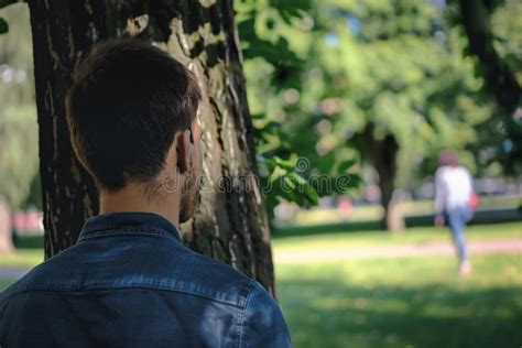 Back View Of Man Stalking Woman While Hiding Behind Tree In Public Park