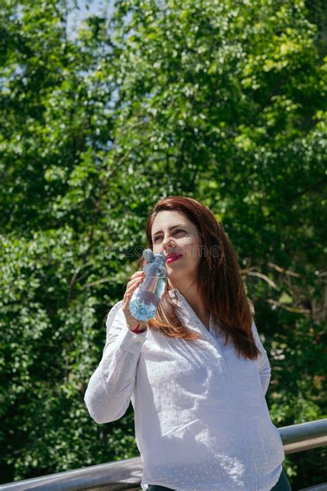 Mature 45 Year Old Woman With Long Hair And Red Lips Drinking Water From A Bottle In Nature On A